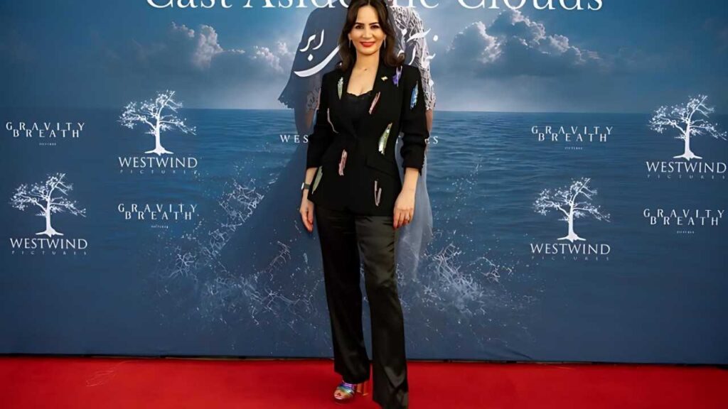 Shohreh Aghdashloo in a black blazer with colorful streaks stands on a red carpet before a blue backdrop with a white tree logo.