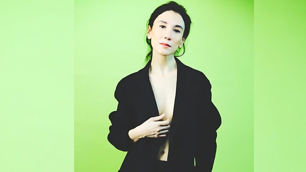 Sibel Kekilli with dark hair in a black blazer against a bright lime-green backdrop, gazing at the camera.