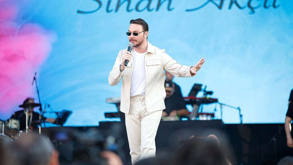 Sinan Akçıl in cream suit and white tee, sunglasses, microphone in hand, performing on blue stage with band equipment behind.