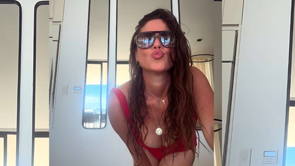 Soleil Moon Frye with wavy hair in a red bikini puckers lips toward the camera, wearing sunglasses in a bright room with ocean view.