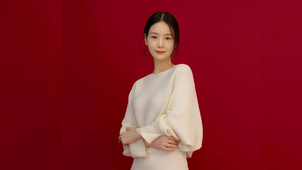 Son Ye-eun in a cream knit sweater with voluminous sleeves stands against a bold red backdrop, arms folded softly, smile.