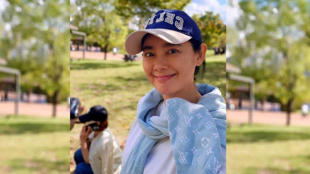 Song Seo-yeon in a light blue LV-pattern scarf and blue cap poses in a sunlit park, with a blurred person in the background.