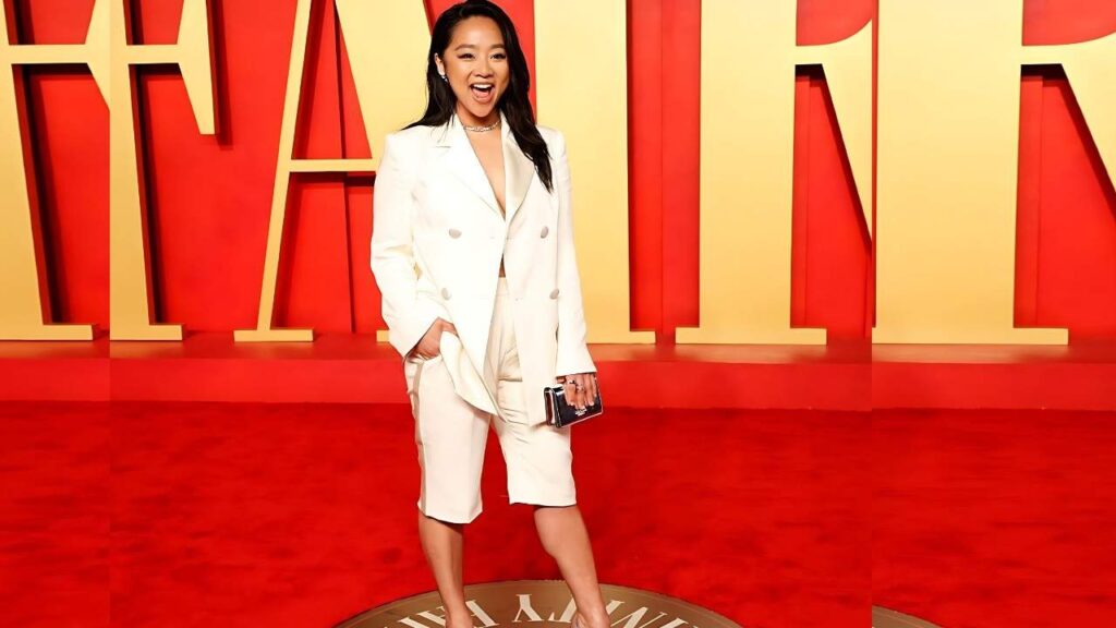 Stephanie Hsu in a white blazer and knee-length shorts poses on a red carpet, clutch in hand, with gold letters behind.