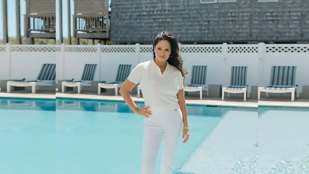 Tanya Memme in white polo and pants poses at a sunny poolside, hand on hip, lounge chairs and white fence behind.