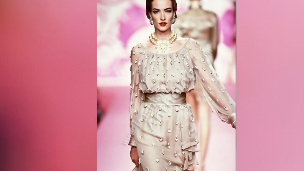 Tatjana Patitz walks a pink runway in an ivory sheer dress dotted with pearl beads, cinched at the waist with ruffled layers and a bold pearl necklace.