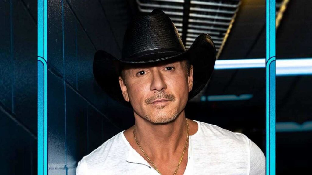 Tim McGraw in a black cowboy hat and white shirt with a gold chain, standing in a blue-framed hallway backdrop.