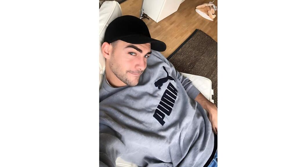 Tom Austen in a gray PUMA sweatshirt and black cap, smiling at the camera on a wooden floor