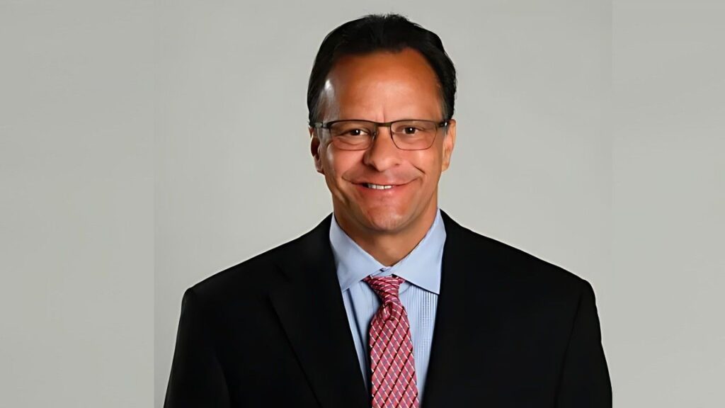 Tom Crean in a dark suit, light blue shirt, and pink patterned tie, wearing glasses, against a neutral gray backdrop.