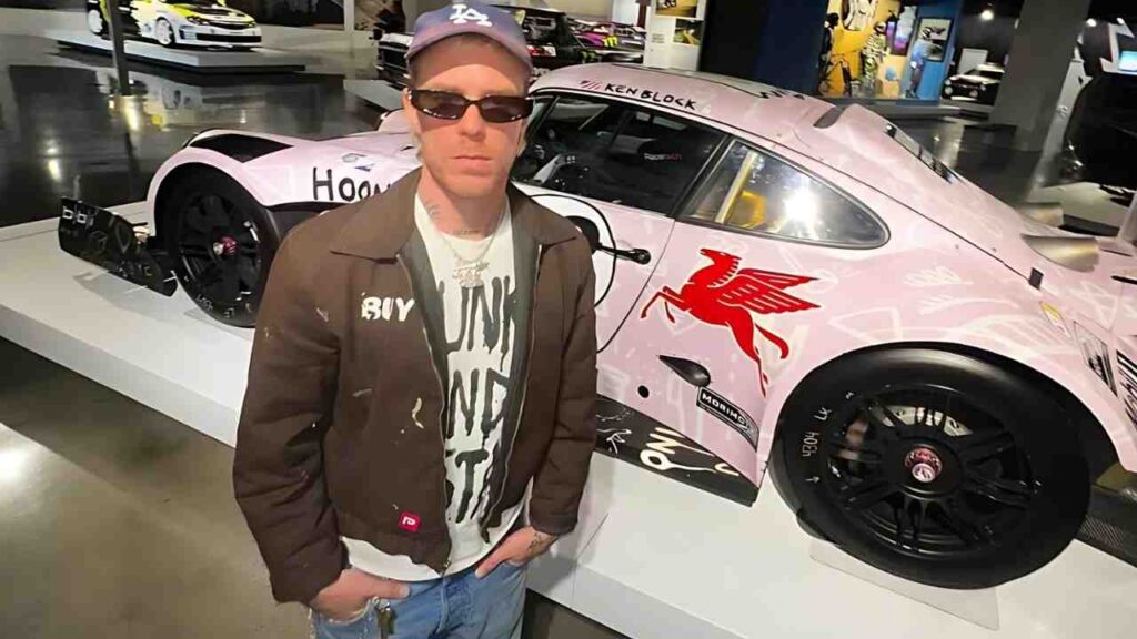 Trevor Andrew in a brown jacket, white graphic tee, cap and sunglasses stands beside a pink racing car with a red Pegasus emblem in a showroom.
