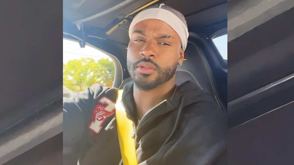 Trevor Jackson in a black jacket and white headband sits in a car, yellow seatbelt across his chest, gazing at the camera.