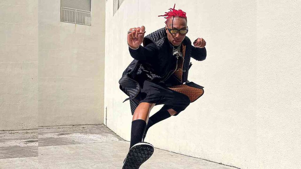 Tyla Yaweh with red hair and sunglasses leaps beside a wall, wearing a black jacket, shorts, socks, and a tan bag.