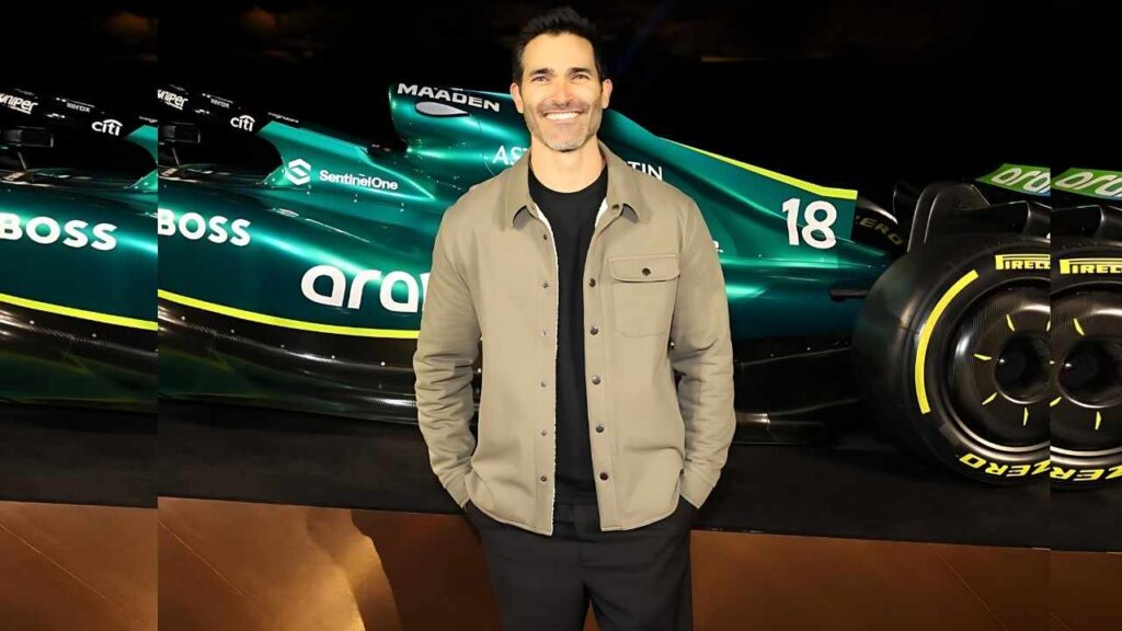 Tyler Hoechlin in a beige jacket and black shirt stands with hands in pockets before a teal Formula 1 car with yellow trim and the number 18.