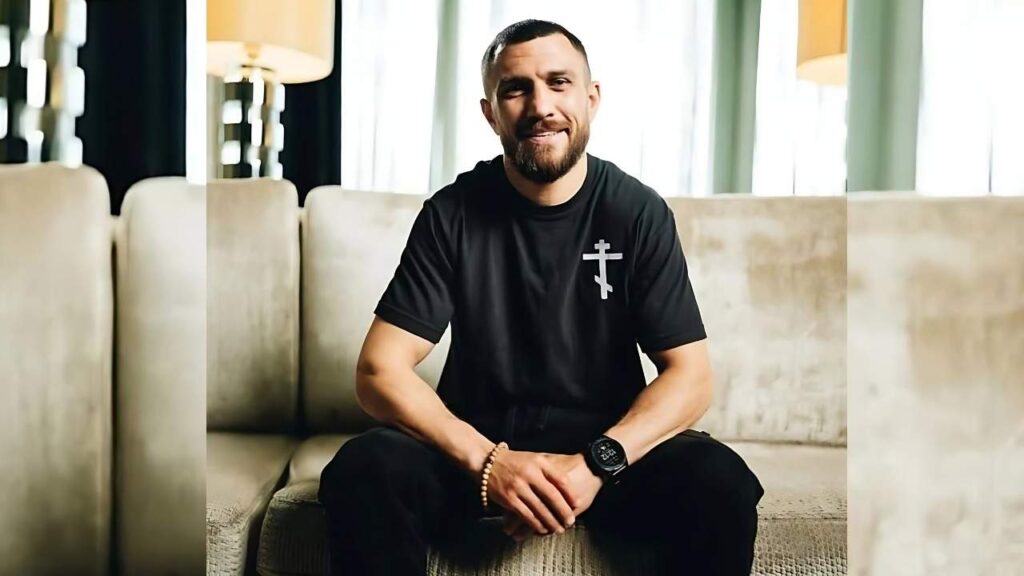 Vasyl Lomachenko with a beard in a black T-shirt with a white cross sits on beige sofa, hands clasped, wearing a black watch.