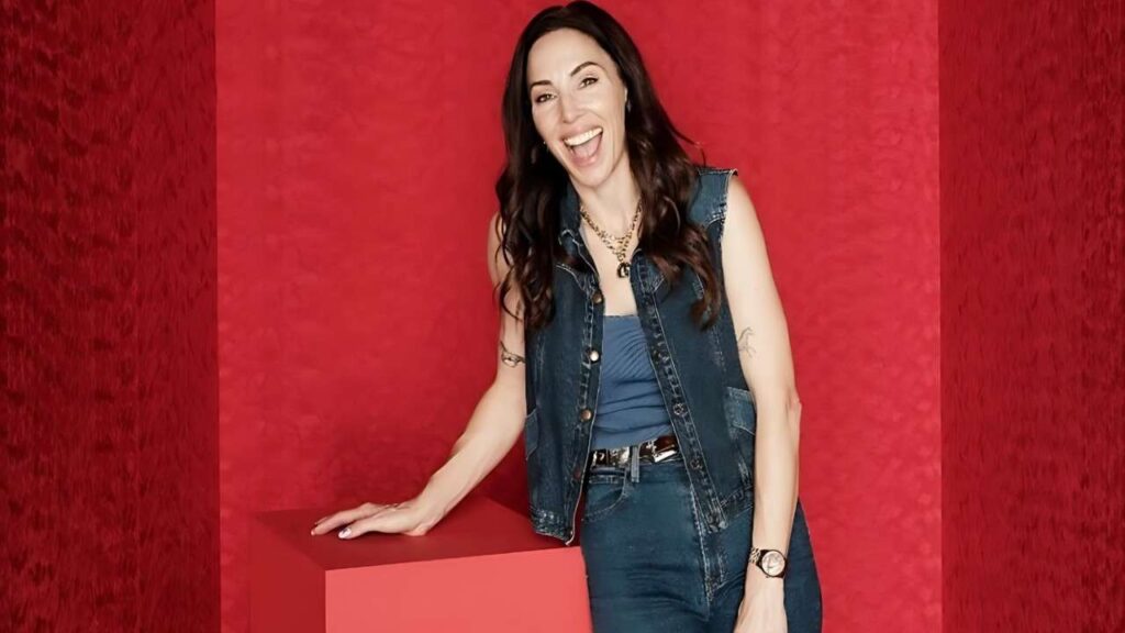 Whitney Cummings with long dark hair in a denim vest and jeans, leaning on a red block against a red-textured background.