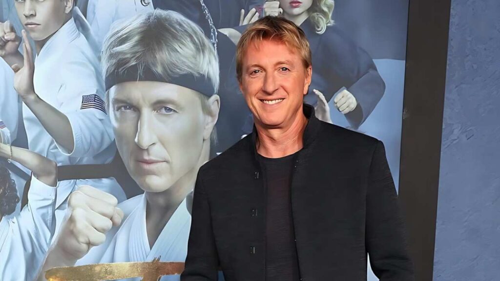 William Zabka in a dark jacket stands before a martial-arts poster showing fighters in white gi and a stern headband portrait.