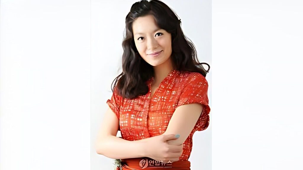 Yeo Woon-kye with shoulder-length black hair in a red patterned blouse, arms crossed and smiling against a white backdrop.