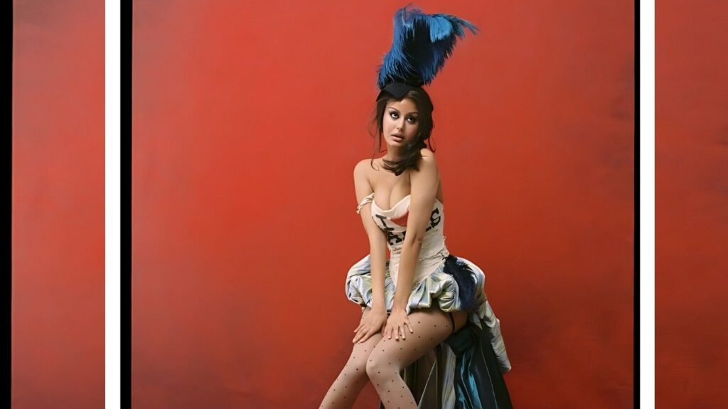 Zahia Dehar in a white corseted dress with a blue feather headdress sits forward, hands on knees, wearing dotted stockings against a red backdrop.