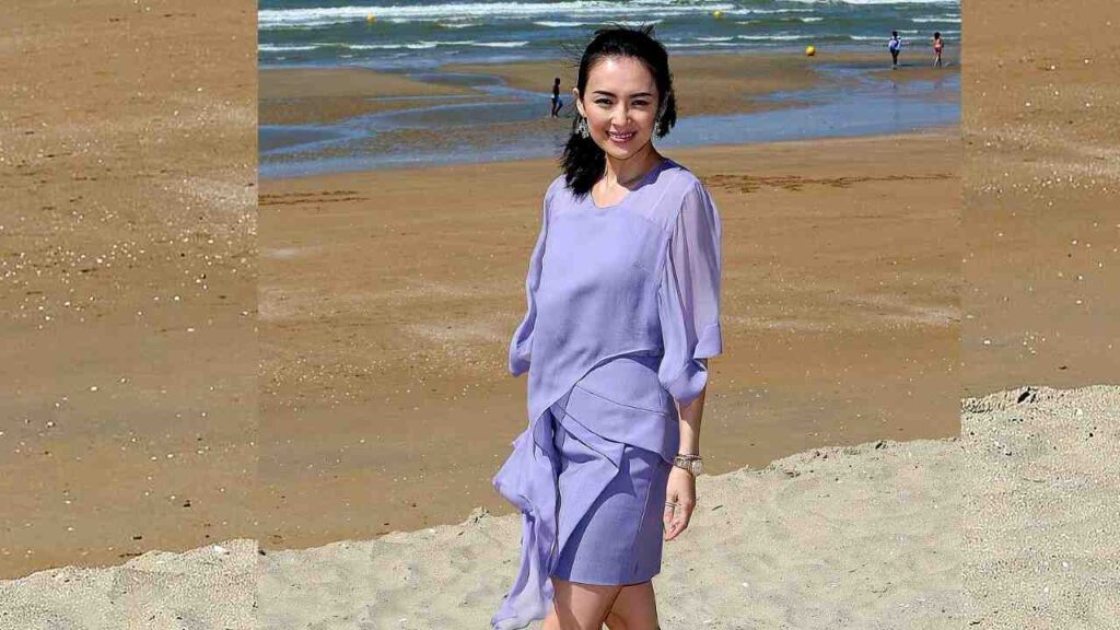 Zhang Ziyi in a lavender knee-length dress with layered wrap-style ruffles stands on a sunny beach, the blue sea behind.
