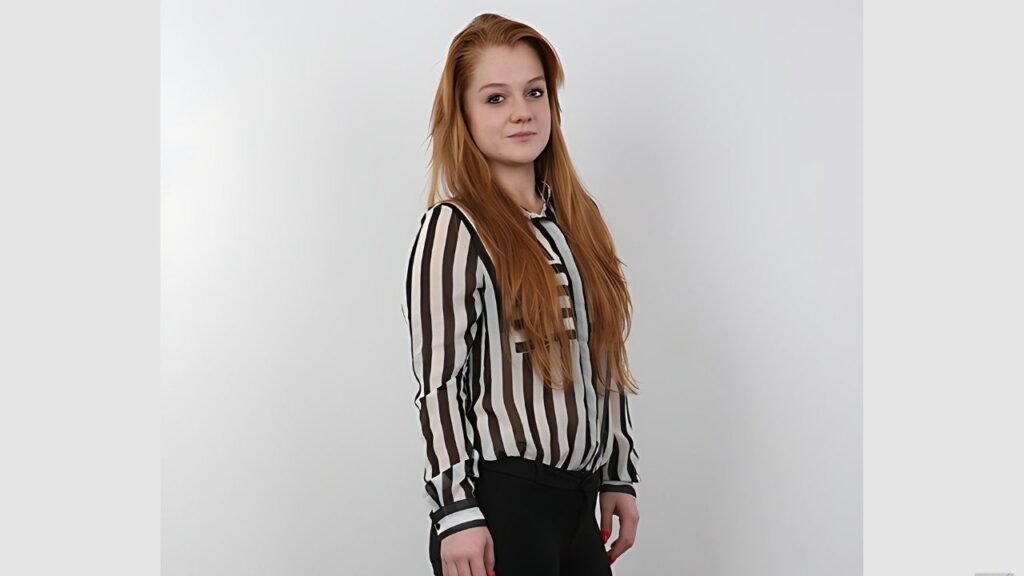 Alex Ginger with long red hair stands against gray backdrop, wearing striped blouse and black pants, gazing to the side.