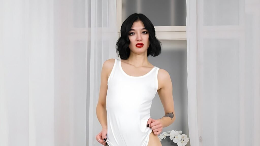 Alex Sen with a black bob, wearing a white tank top, stands before sheer white curtains, red lipstick, holding sides.
