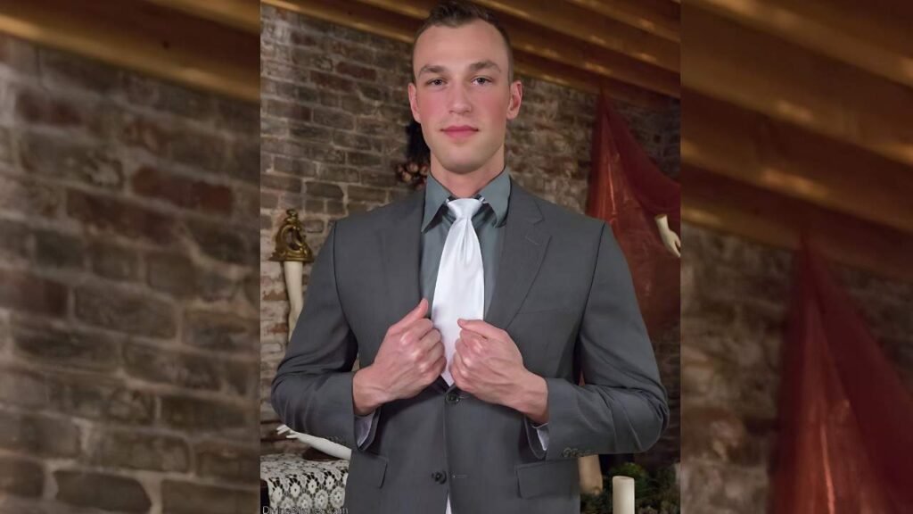 Alrik Angel in a gray suit adjusts his jacket, holding a white tie, against a brick-walled backdrop with draped decor.