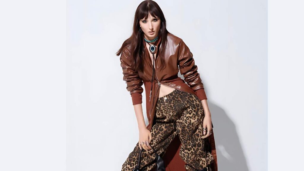 Alyssa Moore with long dark hair and bangs wears a brown leather bomber jacket over leopard-print pants, turquoise choker, posing against a pale backdrop.