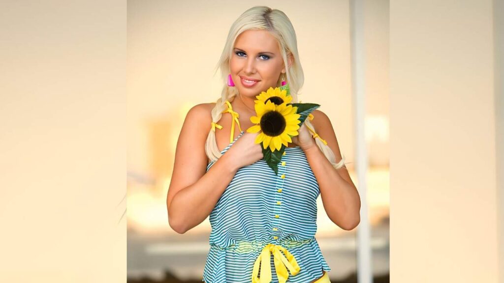 Amy Varela with twin braids and pink earrings holds a sunflower bouquet, wearing a blue striped top with a yellow waist tie.