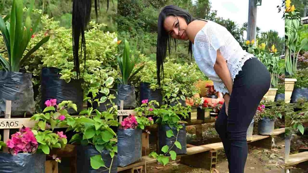 Andrea Flores in white lace top and black pants bends over potted plants at an outdoor garden shop, pink flowers in foreground.
