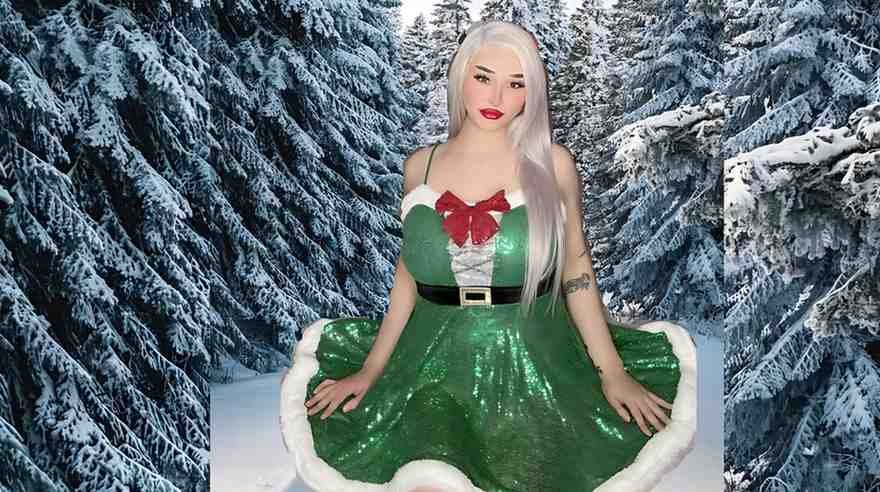 Angel Ducati with long platinum hair sits in a sparkly green elf dress with white fur trim, red bow, and belt, in a snowy forest.