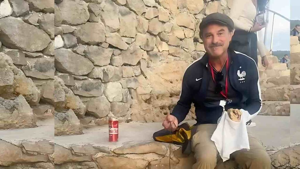 Anthony Crane wearing a dark track jacket and flat cap sits on a rough stone wall, with a Coca-Cola can, napkin and food nearby.