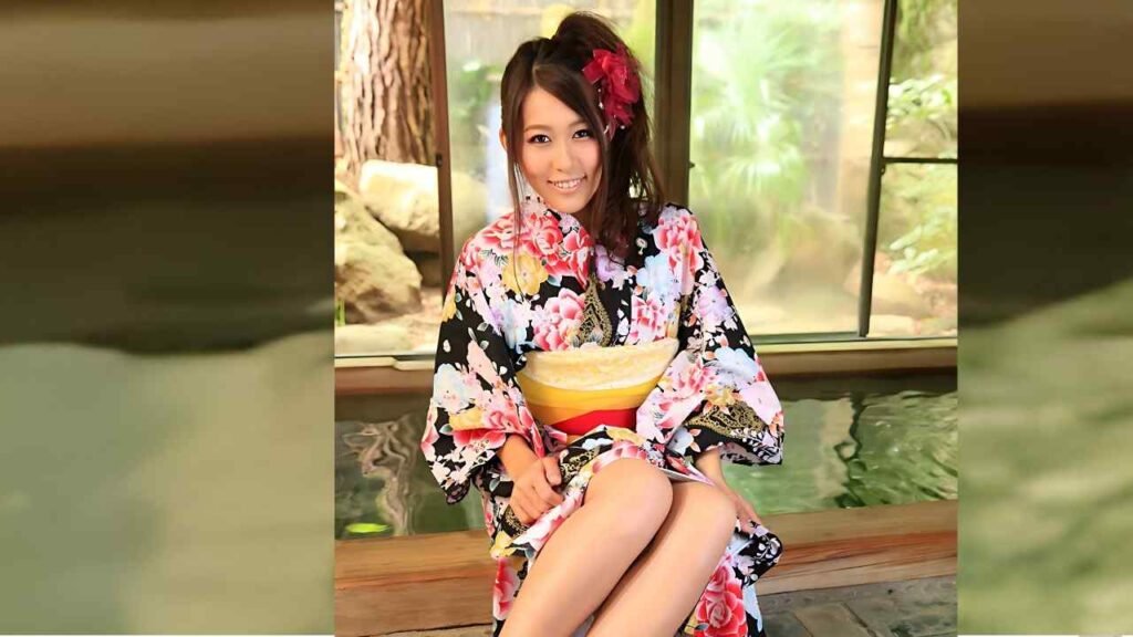 Aoi Mizuno (水野葵) in a bright floral kimono sits on a wooden bench by a window, red hair flower, pastel obi, garden view.