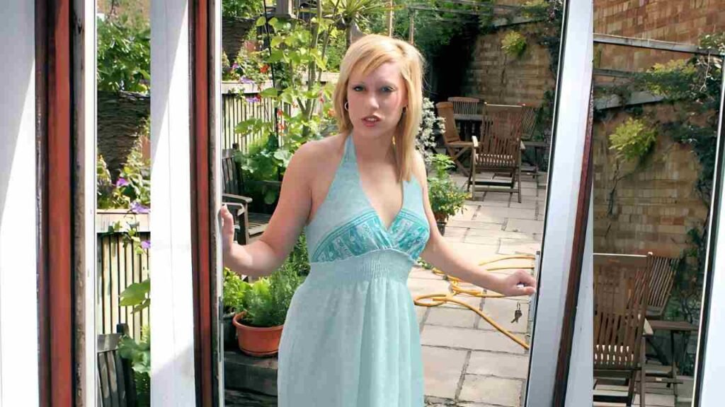 Axa jay in a light blue halter dress stands in an open doorway to a sunlit garden patio, hands on the frame.