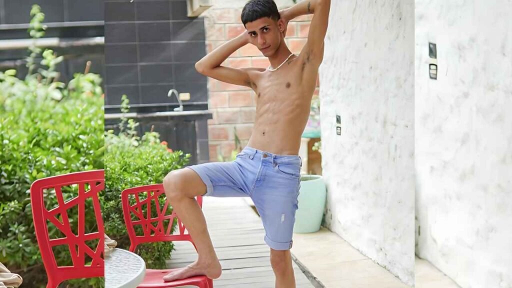 Axel Turner in light blue denim shorts poses with hands behind his head on an outdoor deck, one foot on a red chair beside white marble wall.