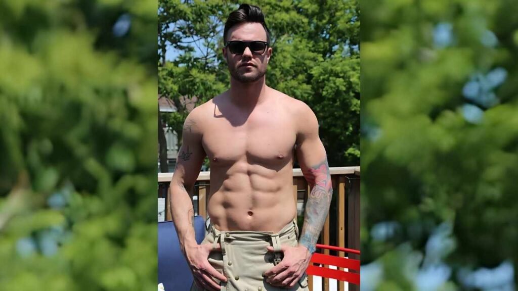 Beau Reed with sunglasses poses on a sunlit deck, hands on beige khaki shorts, muscular torso, tattoos on arms, green trees behind.