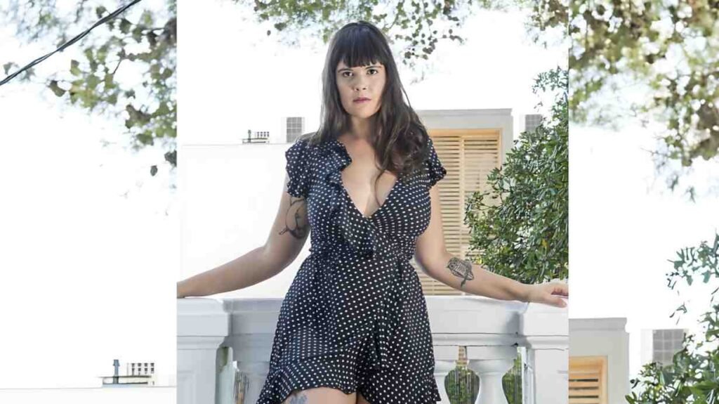 Bel Gris with dark hair and bangs stands on a white balcony in a navy polka-dot dress with ruffled sleeves; arm tattoos visible.