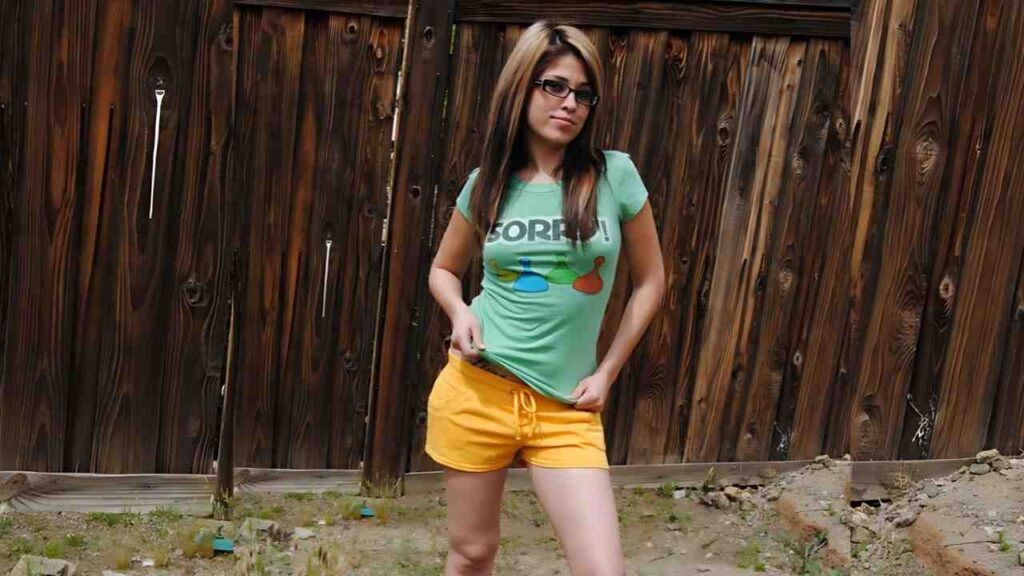 Breana Tabu with glasses, long highlighted hair, wearing a green 'SORRY!' tee and orange shorts, posing casually by a weathered wooden fence.