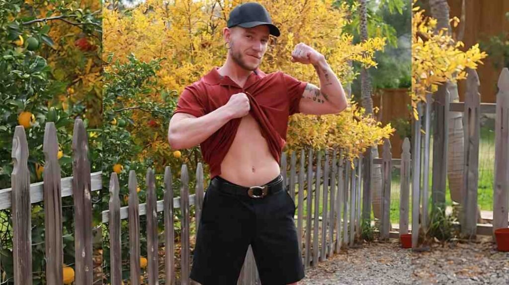 Bruce Jones in red polo and black cap flexes a bicep, lifting his shirt to reveal abs beside a wooden picket fence on a gravel path.