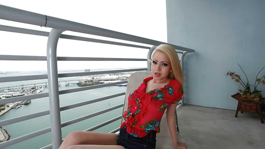 Cami Miller in a bright red floral blouse lounges on a balcony railing, overlooking a marina with boats