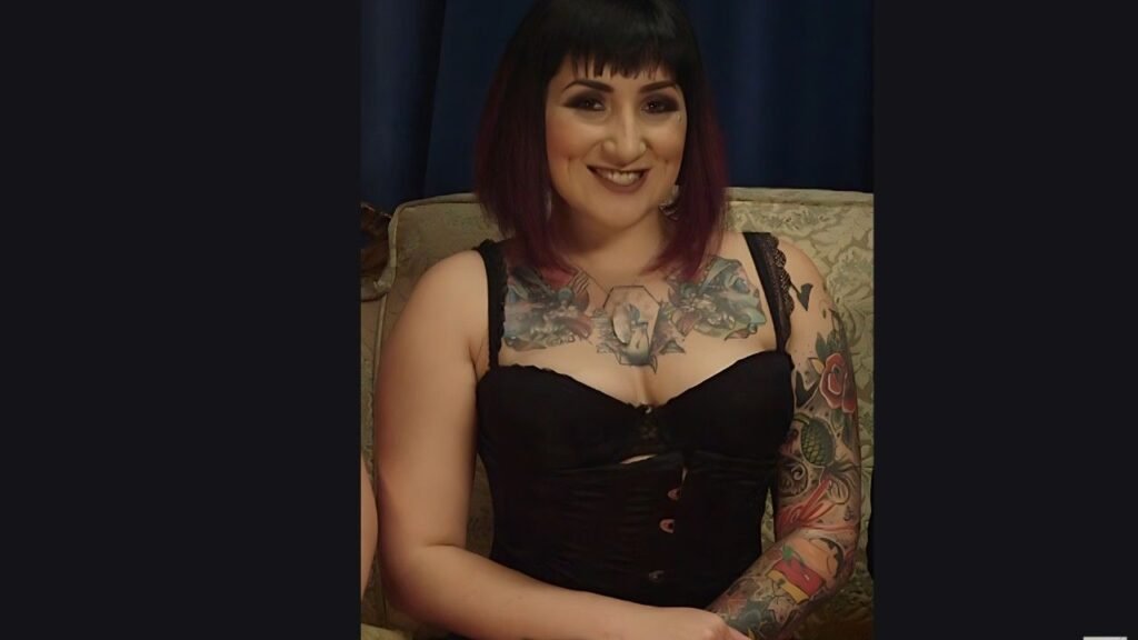 Camille Black with short black-to-purple hair sits on a sofa, wearing a black corset, with tattoos on her chest and arms.