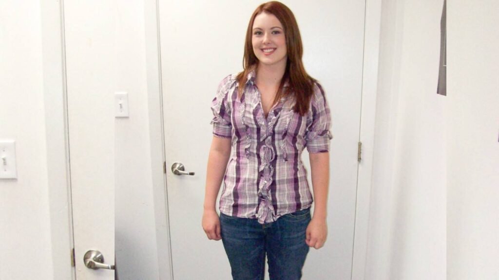 Cammie Fox with long brown hair stands in a bright white hallway, wearing a purple plaid shirt and jeans.