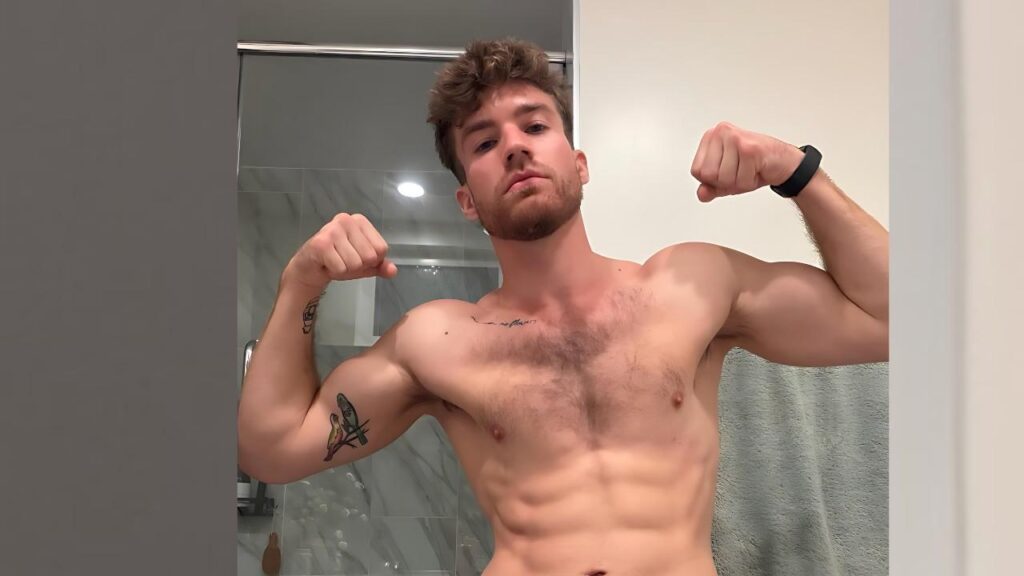 Carter Collins in bathroom mirror, tattoos on left arm, wearing a black wristband; glass shower behind.