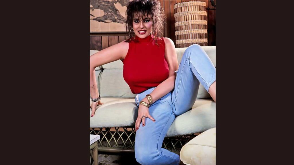 Cathy Patrick in a red sleeveless top and light jeans lounges on a sofa, one leg up, wearing gold bracelets and earrings.