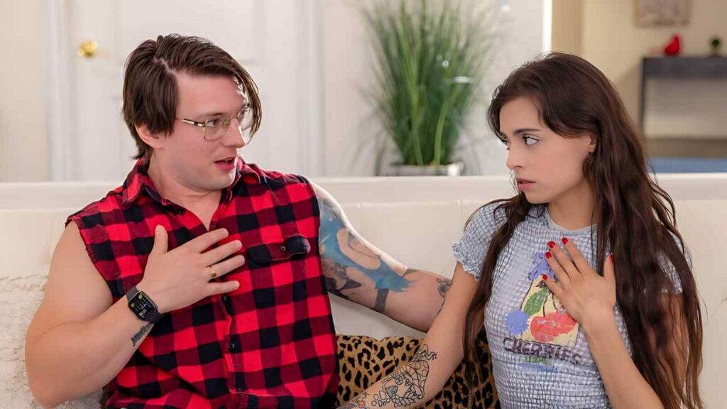 Celtic Iron in red plaid sleeveless shirt with glasses talks to a surprised woman with long dark hair and a floral top; both hands on their chests on white couch.