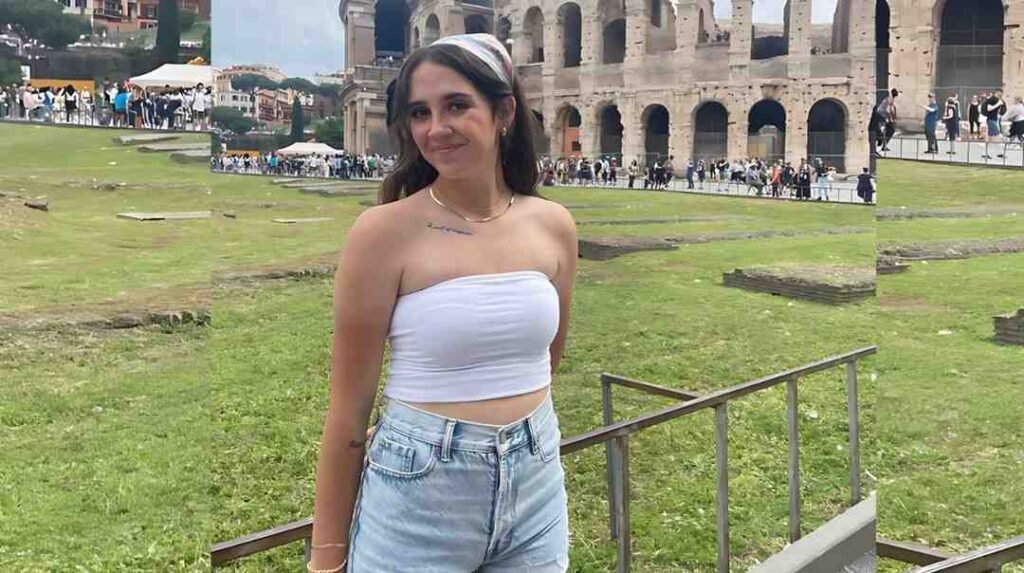 Cheyenne Leigh in a white strapless top and light denim shorts stands on grass by a railing, with Colosseum in the background.