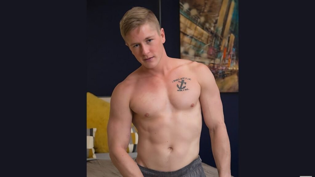 Chris Blades poses in a dark blue room, chest tattoo of an anchor, gray shorts, by a bed with yellow pillows and abstract art.