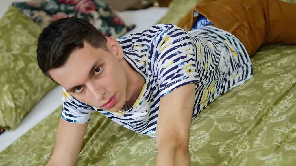 Darin Boswell lies on his stomach on a green floral bedspread, looking at camera in a striped shirt with yellow paint splatters.