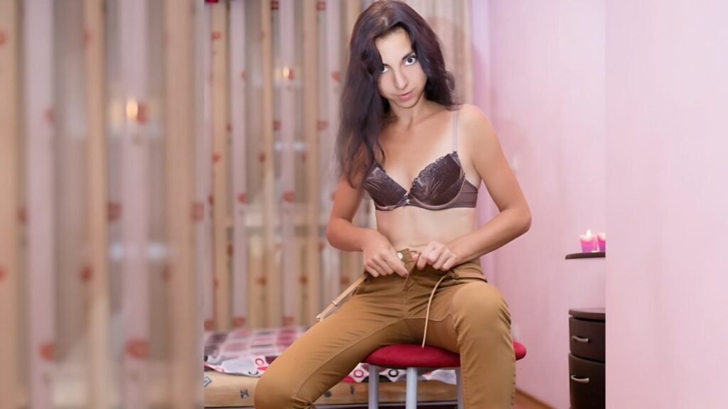 Deja Move sits on a red stool in a room, wearing a bra and tan pants, adjusting a belt; candlelit nightstand nearby.