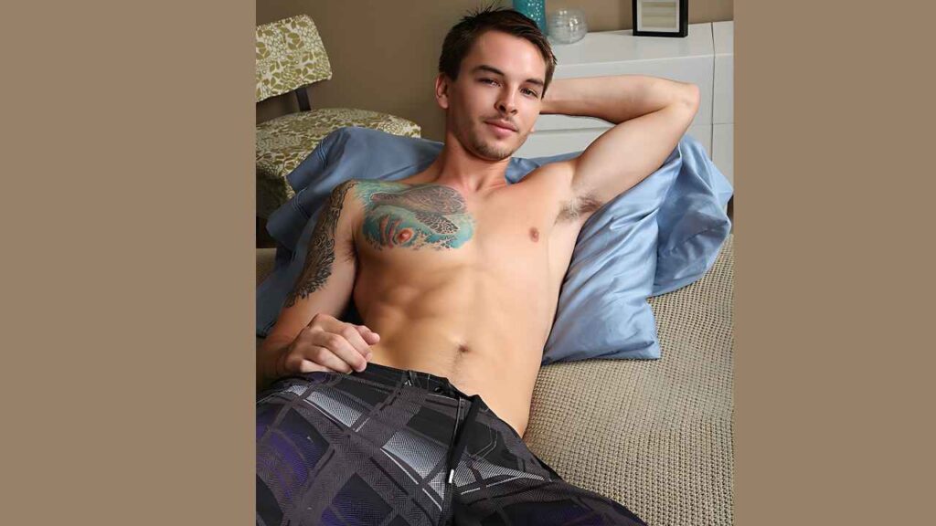 Dorian Ferro lounging on a beige sofa, arm behind his head, colorful chest and left-arm tattoos, wearing dark plaid shorts.