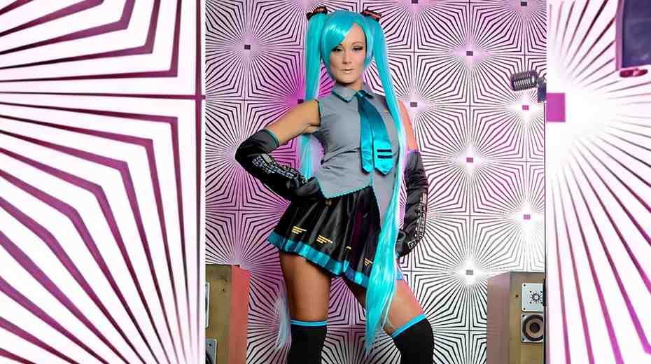 Elizabeth Bally in Hatsune Miku-inspired outfit poses with hands on hips in a pink geometric room; turquoise tails, teal tie.
