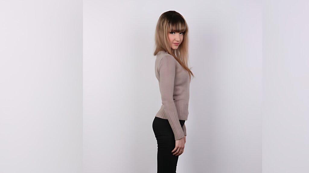 Elly Justin with blonde hair and bangs, wearing a beige knit sweater and black pants against a white backdrop.